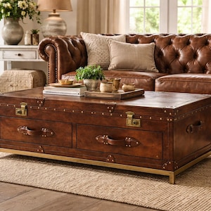 Op de afbeelding: Een grote, rechthoekige, bruin lederen koffietafel in kofferstijl met messing hardware en een goudkleurige basis. De tafel heeft twee lades met leren handgrepen en staat in een woonkamer met een bruine leren bank en een neutraal tapijt.