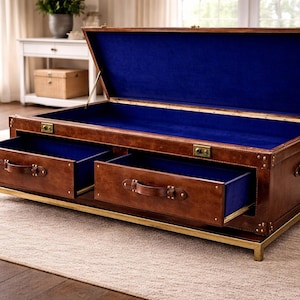 Op de afbeelding: Een bruine leren koffietafel in kofferstijl met twee lades en een blauw interieur. De koffer heeft messing hardware en staat op een goudkleurig metalen frame. Deksel van de koffer is open en onthult het blauwe interieur.
