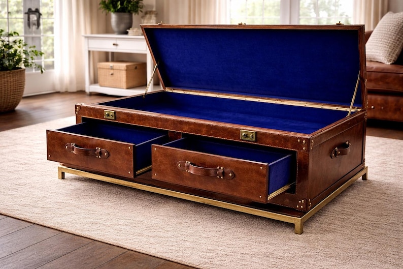 Op de afbeelding: Een bruine leren koffietafel in kofferstijl met twee lades en een blauw interieur. De koffer heeft messing hardware en staat op een goudkleurig metalen frame. Deksel van de koffer is open en onthult het blauwe interieur.