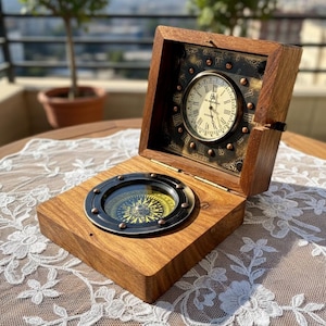 Könnte beinhalten: Eine Holzkiste mit Klappdeckel, mit einer Uhr und einem Kompass. Die Uhr hat ein cremefarbenes Zifferblatt mit römischen Ziffern und einer schwarzen Lünette. Der Kompass ist in einem kreisförmigen Rahmen angeordnet. Die Kiste ist aus Holz mit natürlichem Finish.