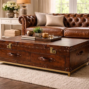 Op de afbeelding: Een bruine leren salontafel in kofferstijl met messing hardware en ladehandgrepen. De tafel heeft een rechthoekige vorm en staat op een goudkleurige basis. De tafel staat in een woonkamer met een bruine leren bank.
