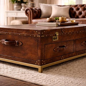 Op de afbeelding: Een rechthoekige bruin lederen salontafel met messing accenten. De tafel heeft twee lades, leren handgrepen en messing hoekbeschermers. Hij staat op een messing frame en staat op een patroonkleed.