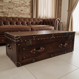 Peut inclure: Une table basse rectangulaire marron foncé de style coffre avec des clous et des poignées de couleur laiton. La table a un couvercle à charnière et un tiroir. La table est devant un canapé en cuir marron. Mesure environ 120 cm de long.