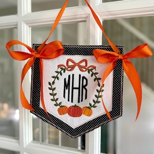 Könnte beinhalten: Ein dekoratives Banner mit einem weißen Mittelteil mit Monogramm und Kürbissen, umrahmt von einem schwarzen Polka-Dot-Rand. Es ist mit orangefarbenen Schleifen und Bändern zum Aufhängen verziert, perfekt für die Herbstdekoration.