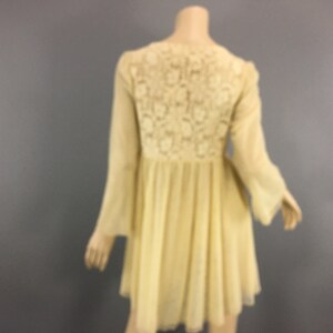 May include: A pale yellow dress with long, flared sleeves and a flowing skirt. The back of the dress has a detailed floral lace panel. The dress is shown on a mannequin against a grey background.