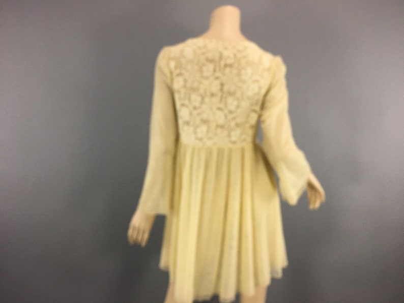 May include: A pale yellow dress with long, flared sleeves and a flowing skirt. The back of the dress has a detailed floral lace panel. The dress is shown on a mannequin against a grey background.