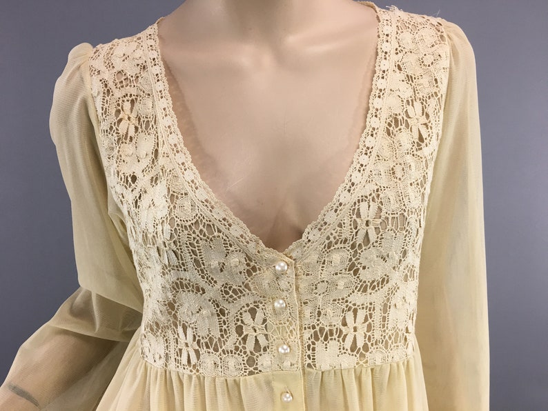 May include: A cream-coloured vintage nightgown with a V-neck and long sleeves. The bodice has detailed lace, with pearl-like buttons down the front. The sheer fabric drapes from the waist, creating a flowing silhouette.