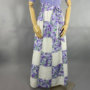 May include: A long white and purple floral skirt with a patchwork design. The skirt has a white eyelet fabric and a purple floral print. The skirt is tied at the waist with a purple ribbon.
