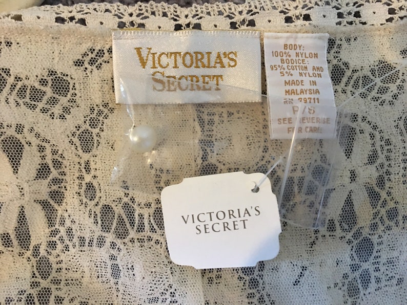 May include: Off-white lace garment with Victoria's Secret tags. The tags display the brand name, material details, and size. The lace features a delicate floral design.