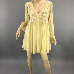 May include: A pale yellow dress with a V-neck and long, flowing sleeves. The bodice features intricate cream-coloured lace detailing. The dress has a button-down front and a flared skirt, creating a bohemian aesthetic. The dress is displayed on a mannequin.