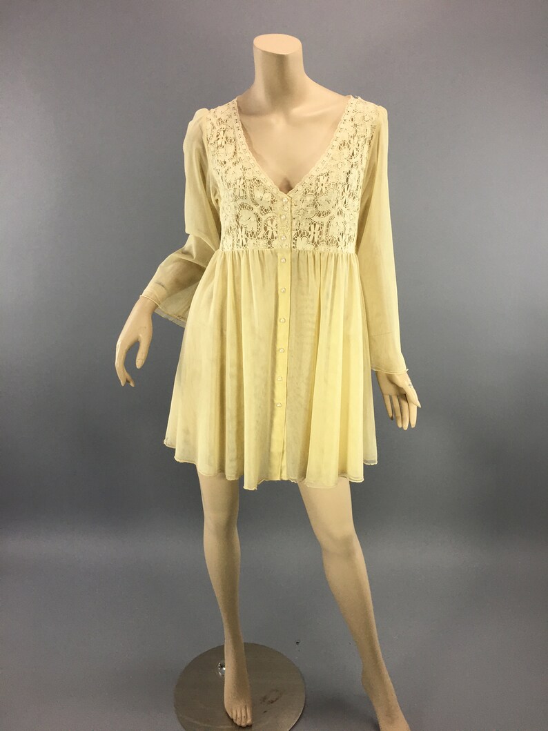 May include: A pale yellow dress with a V-neck and long, flowing sleeves. The bodice features intricate cream-coloured lace detailing. The dress has a button-down front and a flared skirt, creating a bohemian aesthetic. The dress is displayed on a mannequin.