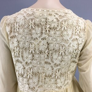 May include: Cream-coloured dress with a detailed lace back panel. The dress has long, sheer sleeves and a flowing skirt. The lace detailing features floral and geometric patterns. The dress appears to be made of lightweight fabric.