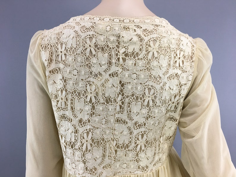 May include: Cream-coloured dress with a detailed lace back panel. The dress has long, sheer sleeves and a flowing skirt. The lace detailing features floral and geometric patterns. The dress appears to be made of lightweight fabric.