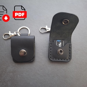May include: Black leather SD card holder with a snap closure and a silver keychain attachment. The holder is shown open and closed, with an SD card visible inside the open holder. The holder is designed to protect and carry SD cards.