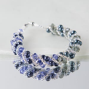 Puede incluir: Pulsera de cuentas con un diseño en espiral. La pulsera está hecha con cuentas azules oscuras y azules claras, con detalles plateados. La pulsera se exhibe sobre una superficie reflectante, mostrando sus intrincados detalles y variaciones de color.