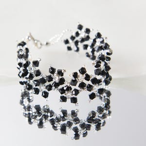 May include: A delicate bracelet featuring a floral design made of small, black, square-cut beads. The bracelet is strung on a silver-colored wire and has a matching clasp. The jewelry is displayed on a reflective surface.