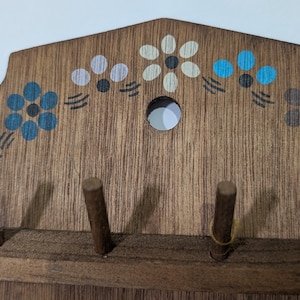 May include: A wall-mounted wooden key holder with a natural brown wood grain finish. The top section features a painted floral pattern in blue, white, and brown. It includes four cylindrical wooden pegs for hanging items and a circular hole.