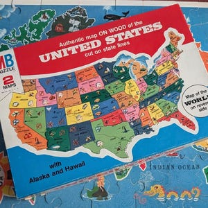 May include: A Milton Bradley puzzle box featuring a colorful map of the United States with text reading Authentic map ON WOOD of the UNITED STATES cut on state lines and with Alaska and Hawaii, resting on a completed world map puzzle.