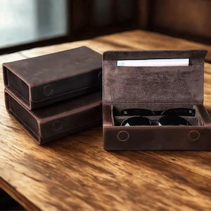 Puede incluir: Un conjunto de estuches de cuero marrón oscuro para gafas. Un estuche está abierto, revelando un forro interior suave de color marrón oscuro y un par de gafas de sol negras en su interior. El exterior tiene un acabado mate con costuras.