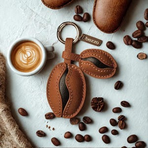 May include: A leather keychain shaped like a coffee bean, with a gold-colored ring and a small tag that reads "bezzo". The keychain is brown with black accents. A cup of coffee with latte art and coffee beans are also in the image.