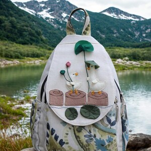 May include: White backpack with a whimsical design featuring two geese with hats and flowers. The backpack has a patterned bottom and side pockets, a top handle, and a zipper closure. The background shows a lake and mountains.