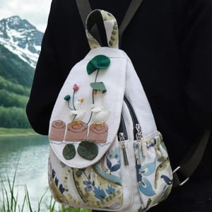 May include: A white backpack with a flap featuring embroidered geese, lotus flowers, and lily pads. The backpack has a patterned side pocket with blue and green floral designs and two zippered compartments. The straps are adjustable.