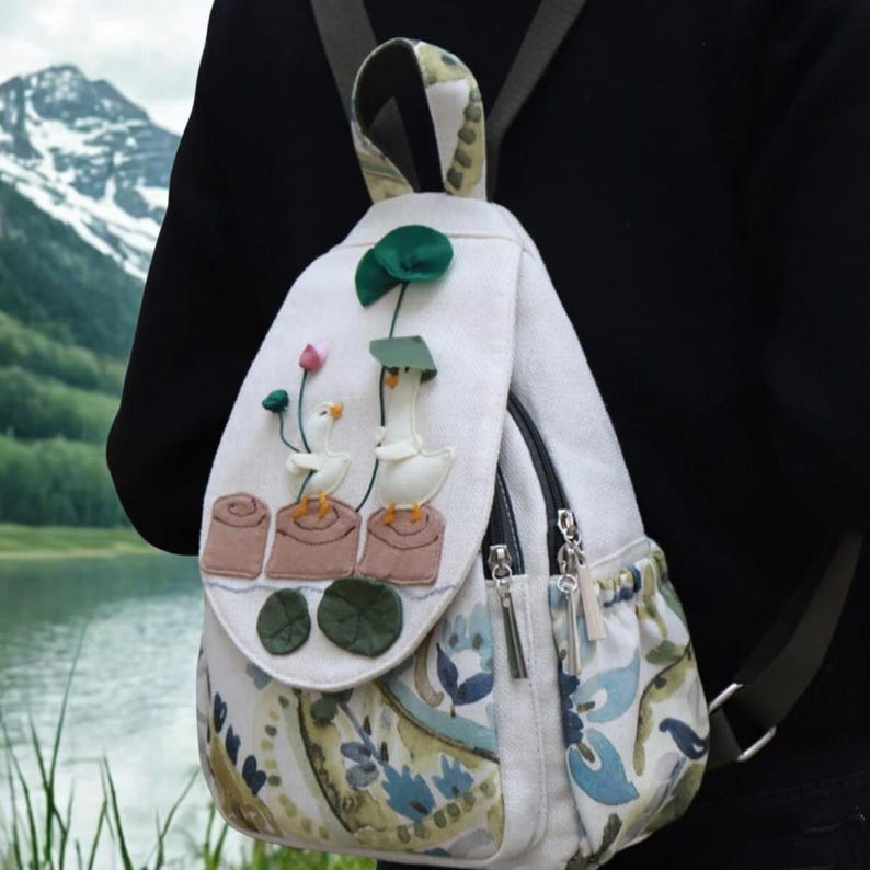 May include: A white backpack with a flap featuring embroidered geese, lotus flowers, and lily pads. The backpack has a patterned side pocket with blue and green floral designs and two zippered compartments. The straps are adjustable.