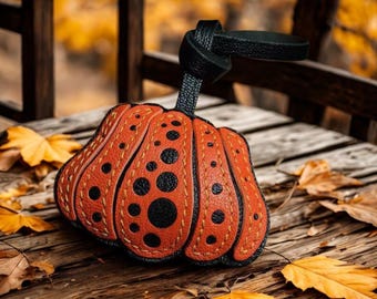 May include: An orange and black leather pumpkin-shaped keychain with black polka dots and a black handle. The keychain is sitting on a wooden surface with fall leaves.