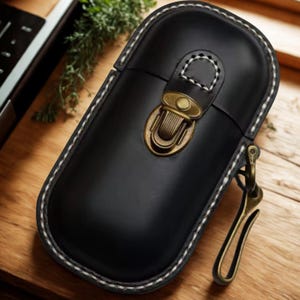 Pode incluir: Estojo de couro preto com costura branca e fecho de latão. O estojo tem formato retangular arredondado e um gancho de latão para prender a um chaveiro ou bolsa. O estojo é provavelmente para guardar itens pequenos.