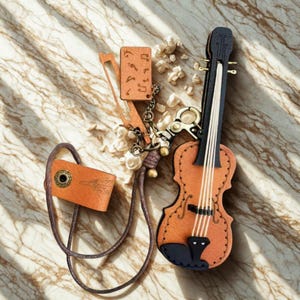 Puede incluir: Un llavero de violín de cuero marrón con detalles negros y cuerdas blancas. Incluye un arco y una pequeña etiqueta de cuero. Está conectado a una correa de cuero marrón.