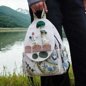 May include: White backpack with a flap featuring appliqued geese, lotus flowers, and lily pads. The backpack has a floral pattern on the bottom and side pockets, with a zippered front pocket and adjustable straps. The background shows a lake and mountains.