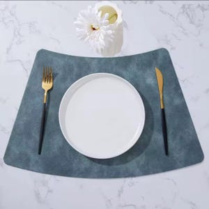 May include: A dining table setting featuring a white plate, gold and black cutlery, and a blue-grey placemat. A white vase with a white flower is in the background. The setting is on a marble surface.