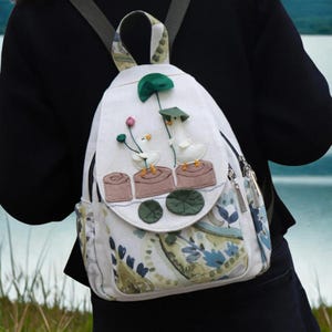May include: White backpack with a whimsical design featuring two geese with hats and flowers. The backpack has a flap with a decorative design and side pockets. The straps and handle are patterned with green and beige.