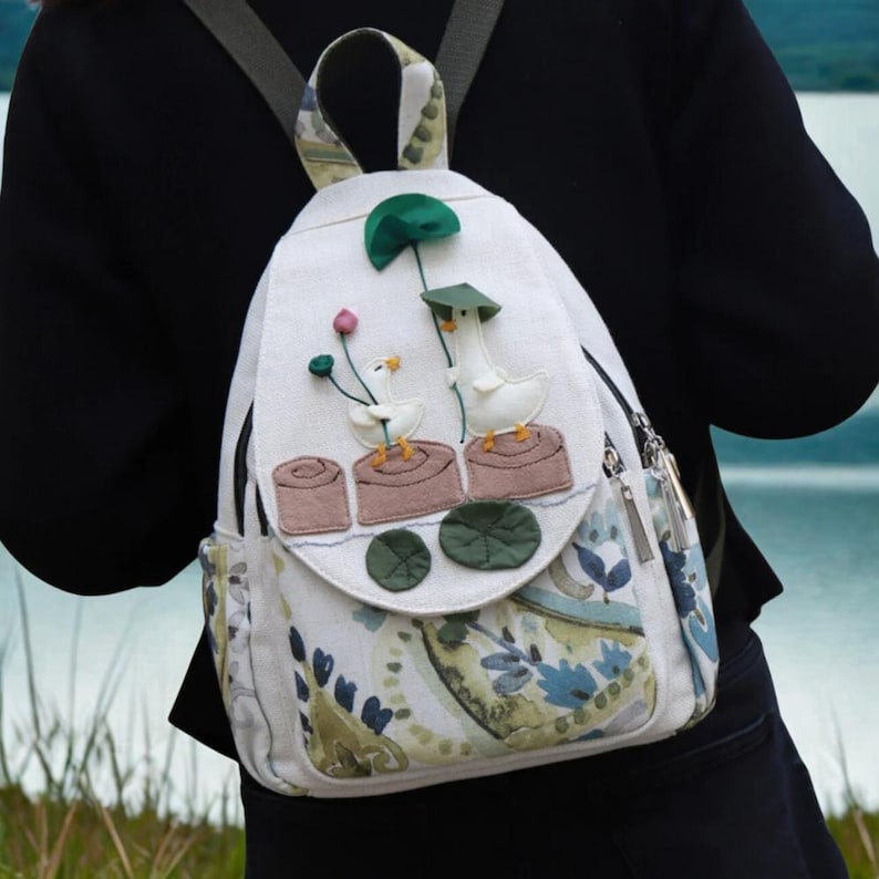 May include: White backpack with a whimsical design featuring two geese with hats and flowers. The backpack has a flap with a decorative design and side pockets. The straps and handle are patterned with green and beige.