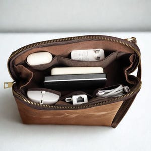 May include: Open brown zippered pouch with multiple compartments, filled with tech accessories. Contents include a white mouse, charging cables, a power adapter, and a small keyboard. The pouch is made of a textured brown material.