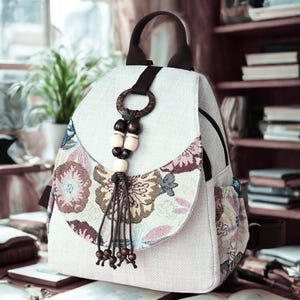 Puede incluir: Mochila beige claro con un estampado floral en la solapa delantera y el bolsillo lateral. La bolsa tiene un asa marrón, un anillo de madera y una borla de cuentas. La mochila está hecha de tela texturizada.