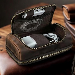 May include: A brown leather travel organiser with a zip closure. Inside, there's a mesh pocket, a white USB charger, and space for cables and a power adapter. The organiser is open, displaying its contents.
