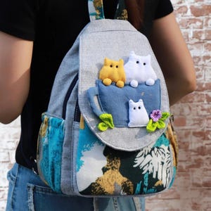 May include: Grey backpack with a blue cup design, featuring three cat figures in yellow and white. The backpack has a colourful patterned front pocket and a zip closure. The straps are patterned with a floral design.