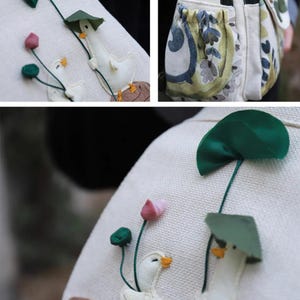 May include: Close-up of a white fabric with embroidered details. Features whimsical designs of white ducks with yellow beaks, wearing green hats, and surrounded by pink and green floral accents. The fabric appears to be part of a larger item, possibly a bag or pillow.