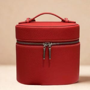 Può includere: Un'accattivante trousse per il trucco cilindrica rossa con una maniglia superiore e una cerniera avvolgente. La trousse presenta cuciture bianche e tiretti della cerniera argentati. Il materiale simile alla pelle ha un aspetto strutturato.