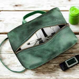 May include: A green leather pouch with a gold zipper, open to reveal white tech accessories and a gray cable. A green strap is attached. A small green container and a black device are in the background.