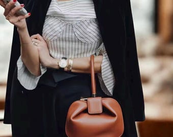 May include: A brown leather handbag with a top handle and a flap closure. The bag is a rounded shape. The person is wearing a black blazer, a striped top, and gold jewelry.