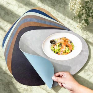 May include: A stack of oval-shaped placemats in various colors, including blue, brown, and gray. A white plate with a salad and a small bowl of dressing sits on top of one of the placemats. A hand is lifting the edge of a blue placemat.