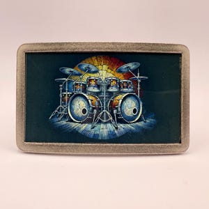 May include: Rectangular metal belt buckle featuring a colorful illustration of a drum kit. The drum set is depicted with blue, brown, and gold tones against a dark green background, with a rainbow-colored backdrop.