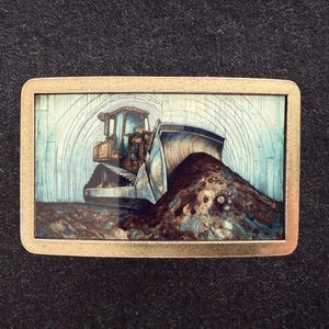 May include: A rectangular belt buckle with a detailed illustration of a bulldozer pushing a pile of dirt. The artwork features shades of brown, blue, and gold, set against a light blue background with arching lines.