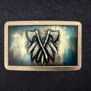 May include: Rectangular metal belt buckle with a detailed graphic of crossed axes and feathers. The design is in shades of gray and white against a blue and teal background, framed by a metallic border. The buckle has a vintage, rustic aesthetic.