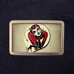 May include: Rectangular metal belt buckle featuring a devil woman design. The image shows a red and black devil woman with horns, a red dress, and black gloves, set against a cream-colored background. The buckle has a silver-colored frame.