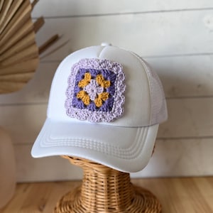 May include: White trucker hat with a crocheted square patch. The patch features a purple border, yellow and orange flower-like shapes, and a light purple center. The hat has a white front and brim, with a mesh back.