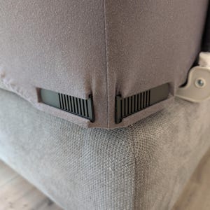 Könnte beinhalten: Nahaufnahme einer grauen Stoffecke, möglicherweise ein Bettlaken oder eine Möbelabdeckung, mit einem schwarzen Lüftungsdetail. Der Stoff ist straff über einer helleren, strukturierten grauen Oberfläche gespannt. Auf der rechten Seite ist ein weiß-grauer Mechanismus zu sehen.