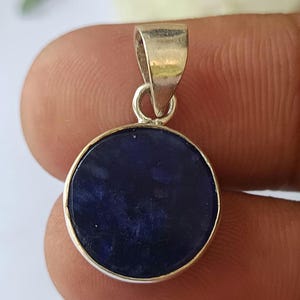 May include: A round, dark blue pendant with a silver-colored bezel and bail. The pendant has a smooth, polished surface and is likely made of a gemstone. The bail is attached to the top of the pendant for easy wear.